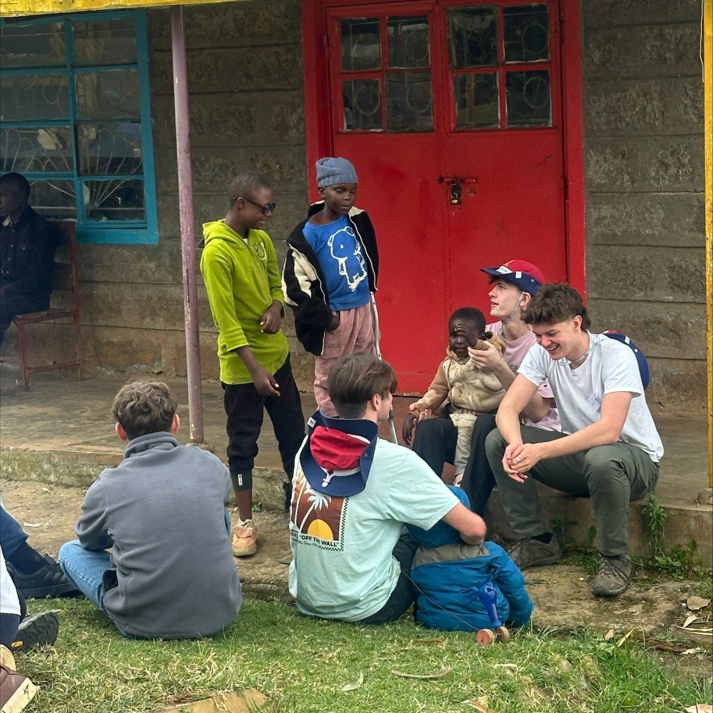 Teenagers and children from Guernsey and Kenya sit and talks outside a stone building with a bright red door and a yellow awning, with others standing nearby.
