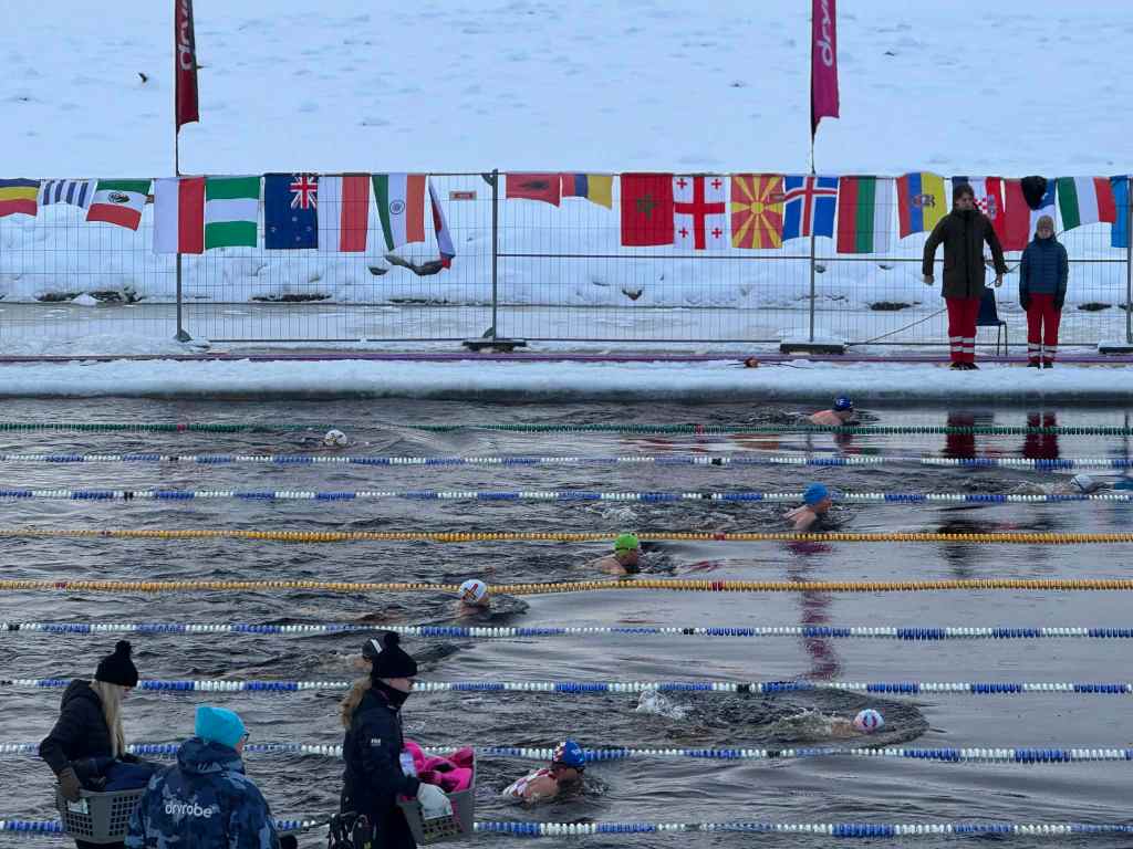 Swimmers in an outdoor ice‑water pool move through marked lanes while flags from various countries hang on a fence behind them.