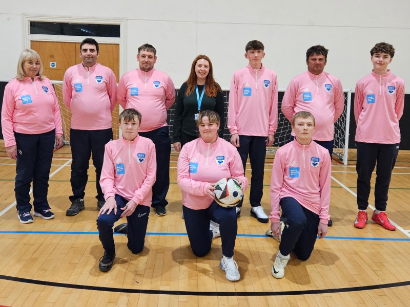 A group of men and women wearing matching pink sports tops pose together in an indoor sports hall, with two people kneeling at the front holding a football.
