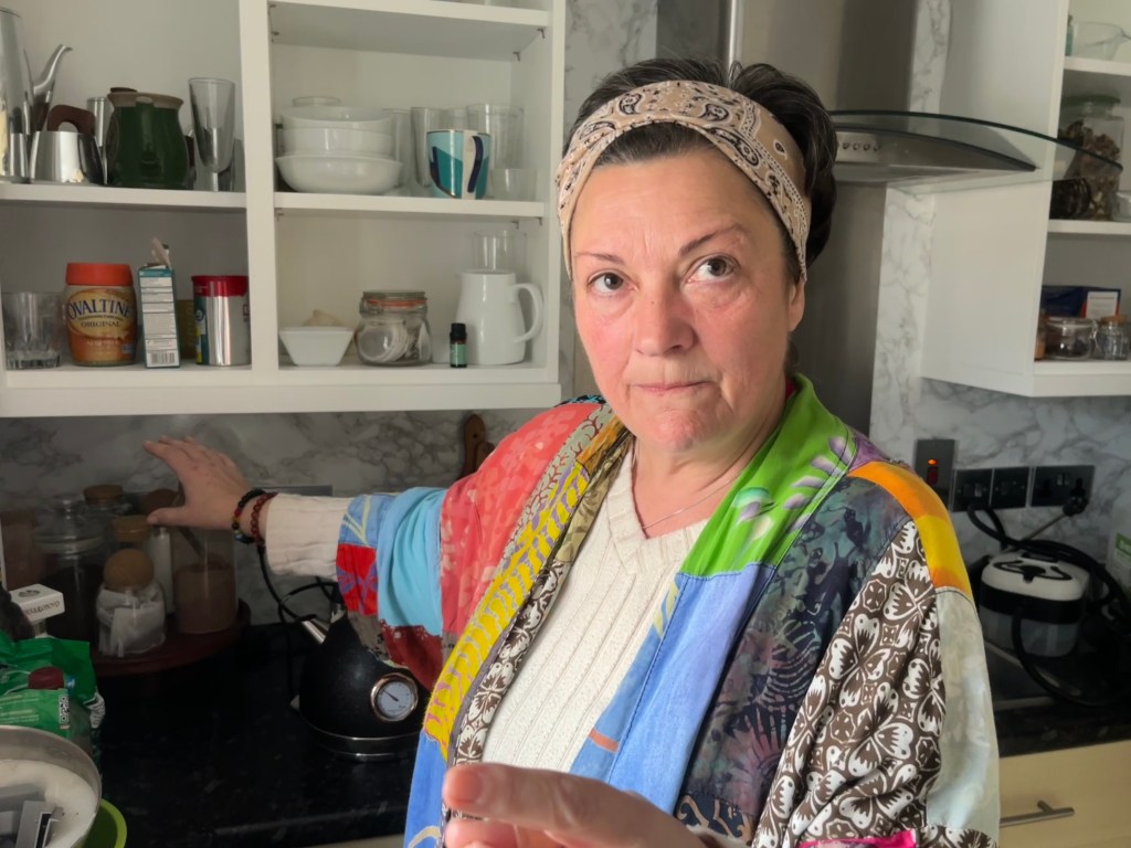A woman standing in a colourful patterned dressing gown and headband stands in a kitchen with open shelves behind her, gesturing toward items on the shelves.