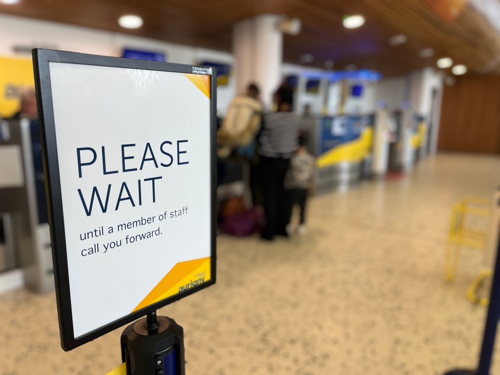 Sign at an airport check‑in area reading ‘Please wait until a member of staff calls you forward.’