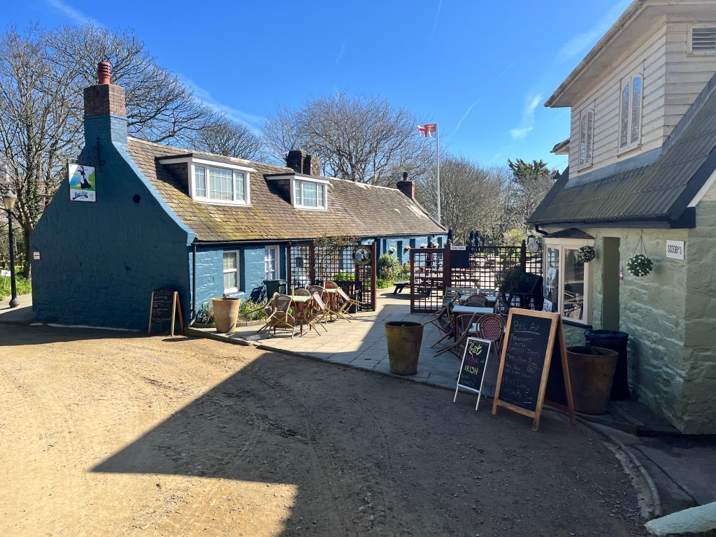 A small cluster of traditional stone buildings - now a pub - along a sunlit dirt lane, including a blue-painted cottage with outdoor tables and chairs on a patio. A sign on one wall shows it's the Bel Air pub. A chalkboard sign and potted plants stand near another light‑coloured building on the right. Leafless trees surround the area, and a flagpole with a red-and-white Sark flag rises in the background under a bright blue sky.