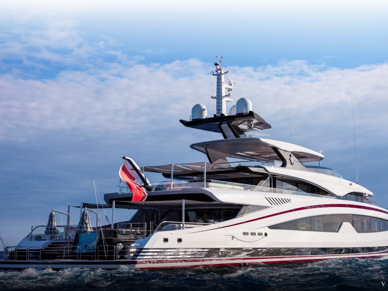 A large, modern motor yacht on open water under a partly cloudy sky. The yacht features multiple decks, sleek white and dark trim, and various antennas on the upper structure. A red-and-white flag is mounted at the stern.