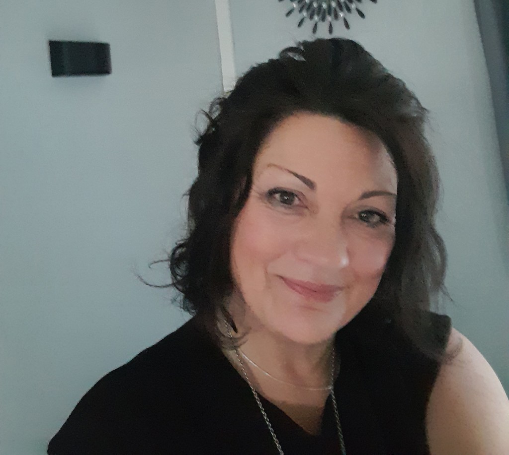 A smiling woman with shoulder-length dark hair in a black top. She's got a necklace on and is in an egg-shell blue living room with a clock on the wall.