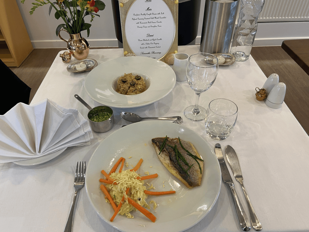 A white table is set for a formal meal with two plated dishes. In the foreground is a large white plate holding a cooked fish fillet garnished with herbs, served alongside shredded vegetables including carrots and cabbage. Behind it is a deep white bowl containing a grain-based dish topped with small round pieces of food. The table includes folded white napkins, cutlery, empty glasses, a small pot of green sauce, a jug, salt and pepper shakers, and a vase of colourful flowers. A decorative printed menu stands upright at the back of the table.