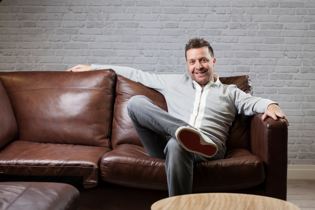 A smiling man sitting casually on a brown leather sofa with one arm resting along the back and one leg crossed over the other. The setting includes a gray brick-pattern wall and a wooden coffee table in the foreground.