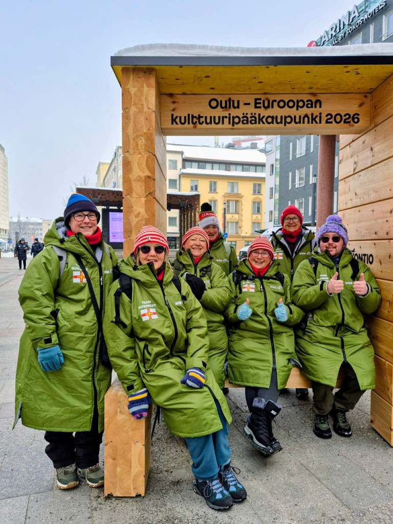A group wearing green winter coats poses at a wooden shelter in Oulu city centre with signage for the 2026 European Capital of Culture.