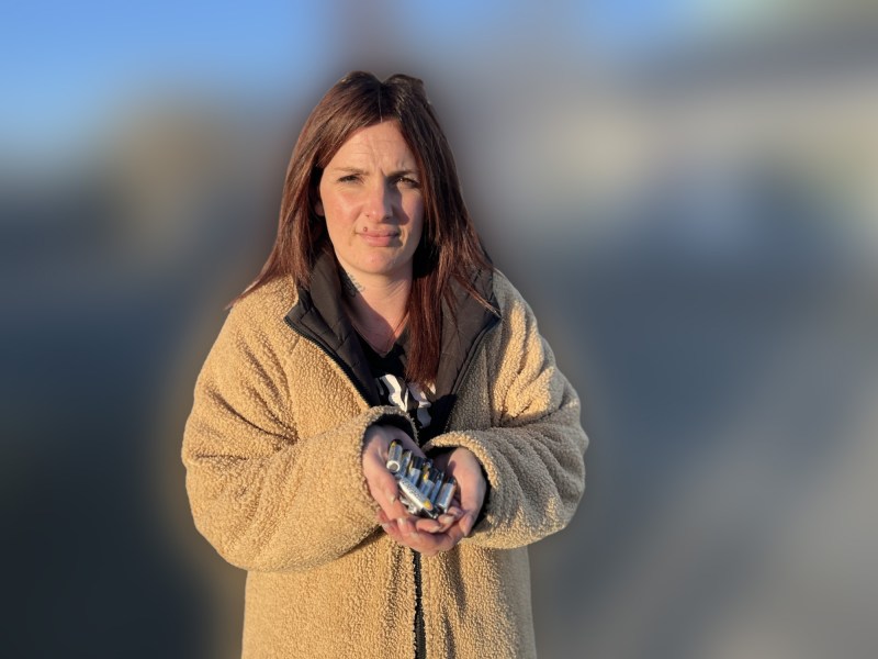 A woman standing in a parking lot at sunset holds a handful of AA batteries toward the camera, wearing a tan fleece jacket, with buildings and parked cars blurred in the background.