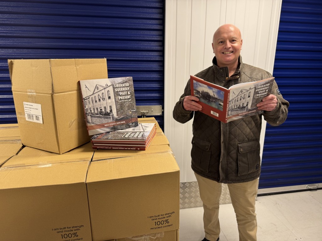 A smiling, bald man in a quilted jacket stands inside a storage unit with blue roller doors, holding open a hardback book titled Liberated Orkneys: Past & Present. Nearby, several cardboard boxes are stacked on a pallet, with more copies of the same book placed on top of the boxes.