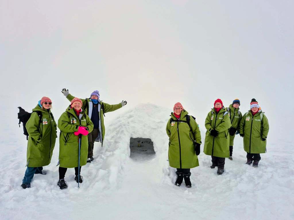 People in matching green winter coats stand on a snowy plain beside a small igloo‑like snow structure in foggy conditions.