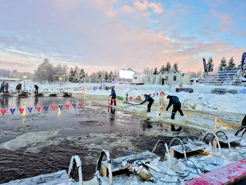 A partly-frozen competition pool with slushy ice in the water as staff clear the surface at sunrise.
