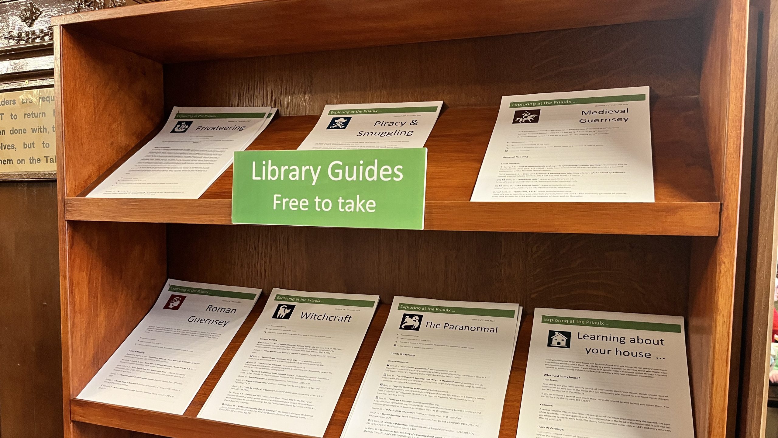 A wooden library display shelf holds several printed information leaflets under a green sign that reads Library Guides – Free to take. The guides include topics such as paranormal phenomena, piracy and smuggling, medieval Guernsey, Roman Guernsey, witchcraft, the paranormal, and researching a house’s history.