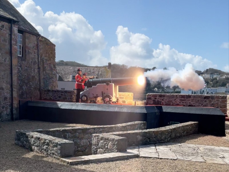 Two heavily bearded men in historic red military-style uniforms fire a cannon from a coastal viewpoint with a large smokey blast and the sea and town skyline in the background.