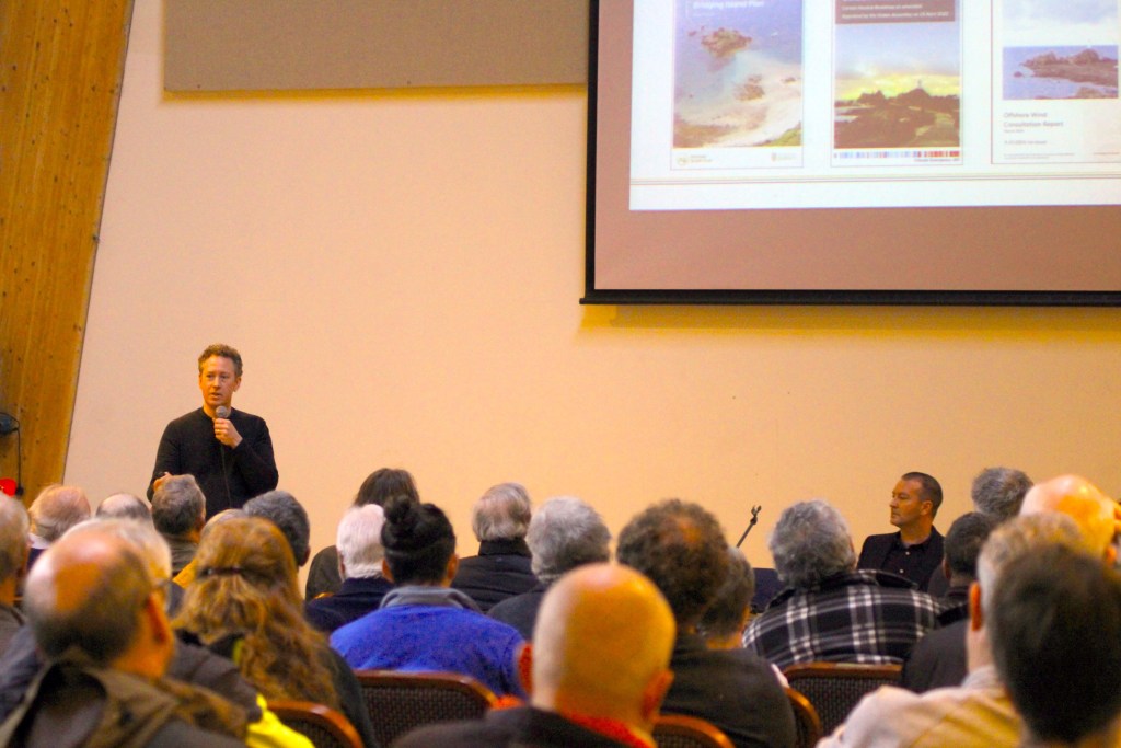 A man speaks into a microphone at the front of a hall while an audience watches a presentation projected on a screen.