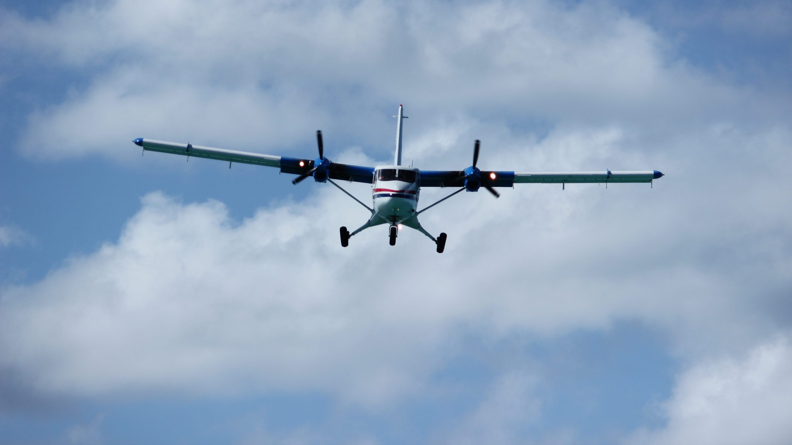 A Twin Otter aircraft flying toward the camera against a partly cloudy sky. It is a small plane with twin propellers.