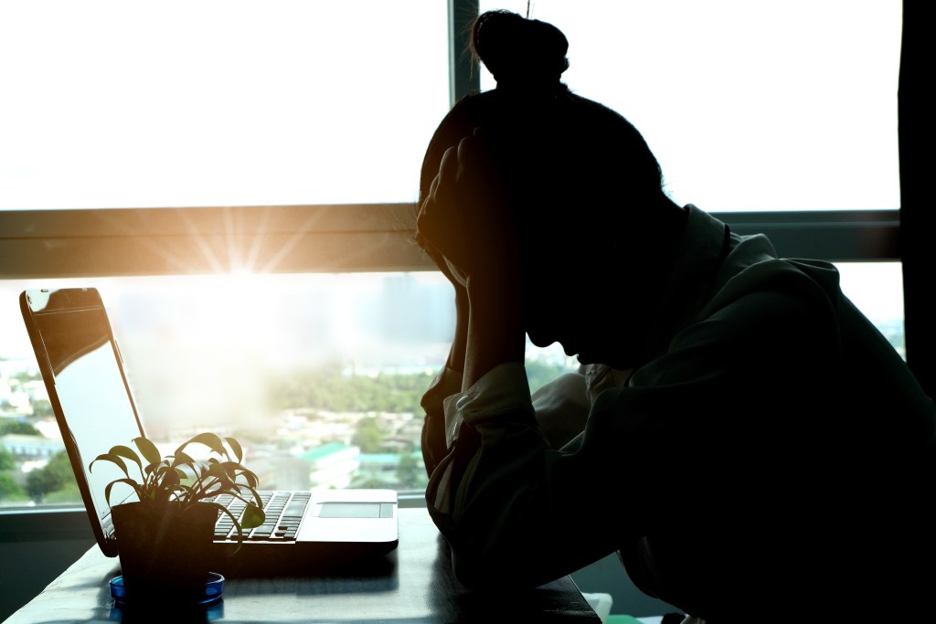 A silhouetted woman sits at a desk with her head in her hands, positioned beside an open laptop. A small potted plant is next to the computer. Bright sunlight shines through a large window in the background, illuminating the landscape outside.