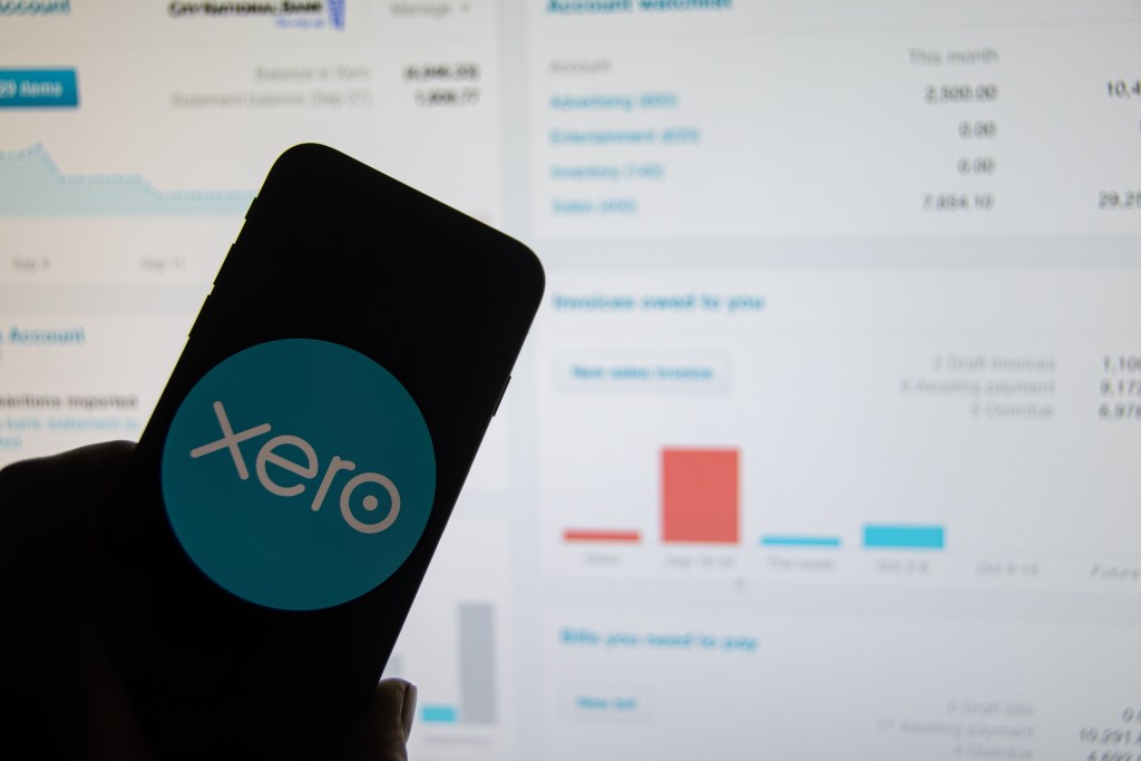 A smartphone displaying the Xero logo is held in front of a computer screen showing financial dashboards and charts.
