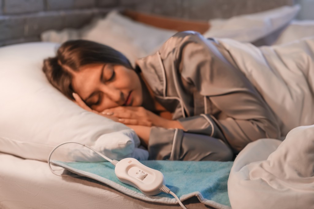 A woman lies on a bed under a duvet, resting on a pillow beside a plugged‑in heating pad with a control unit on top of it.