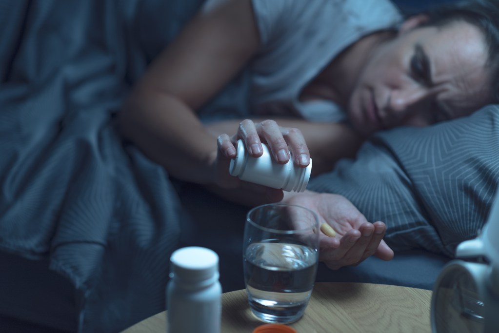 A woman in a dark room lies in bed as she pours a pill out of bottle into her hand - a glass of water close by on her bedside table.