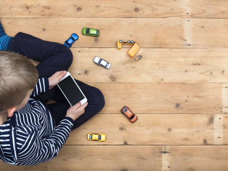 A small boy sits on a wooden floor playing with a mobile phone instead of a collection of toy cars.