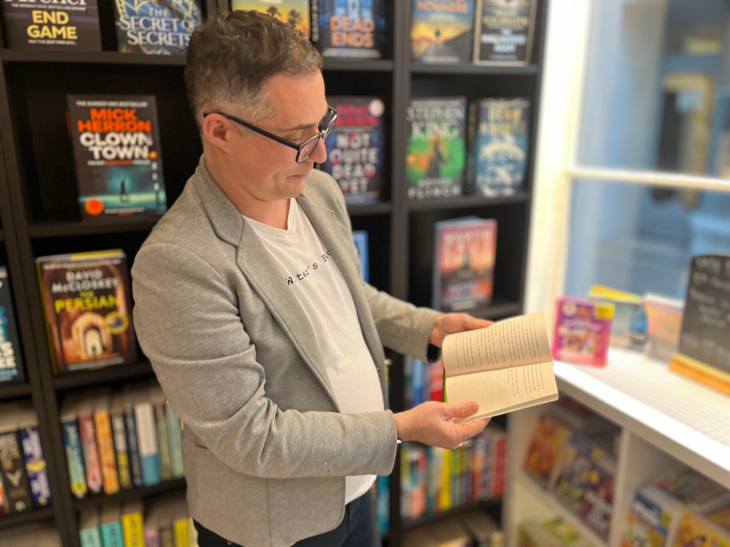 A man in a light-coloured blazer and white T-shirt stands inside a bookstore, holding an open paperback book. Behind the person is a wall of neatly arranged books on dark shelving. To the right, sunlight comes through a window onto a display of smaller items.