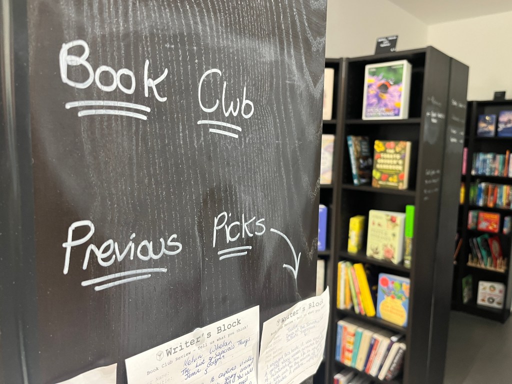 A chalkboard-style panel inside a bookshop displays handwritten text reading Book Club and Previous Picks, with an arrow pointing downward. Below the board are several handwritten recommendation cards titled Writer’s Block, each listing book titles and notes. In the background, tall black bookshelves are filled with colourful books, including children’s and adult titles. The shop is brightly lit, and the shelves extend toward the back of the room.