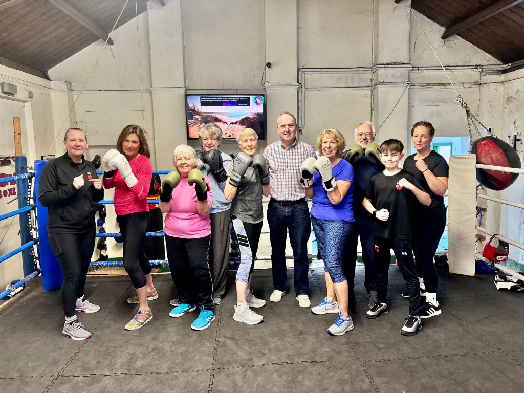 Ten people ranging in age from a small child to pensioners line up in a boxing ring, smiling an posing. Behind them ropes outlining the ring and punchbags.