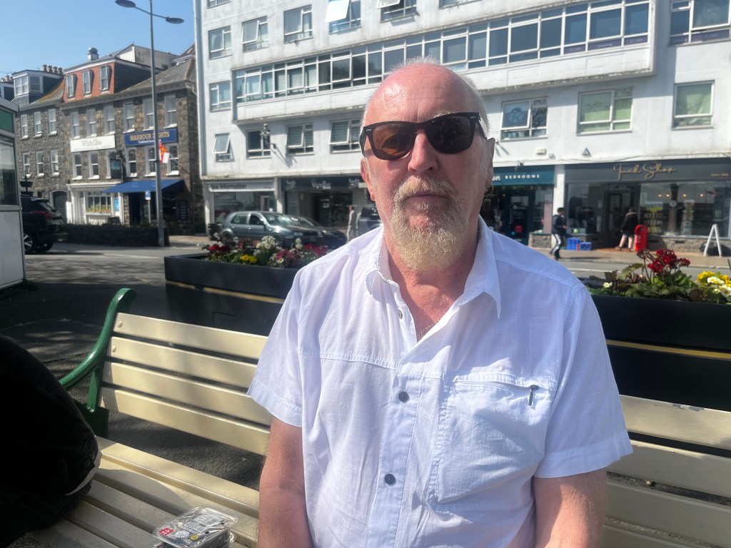 An old, bald man with a grey beard sits on a bench in front of bus stops and a row of shops. He is wearing sunglasses and a short-sleeved white shirt.