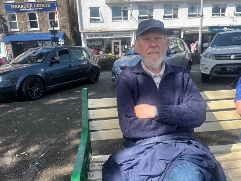 An old man with a grey beard sits on a bench in front of bus stops and a row of shops and cars. He is wearing blue jeans, a blue woollen jumper and a blue cap.