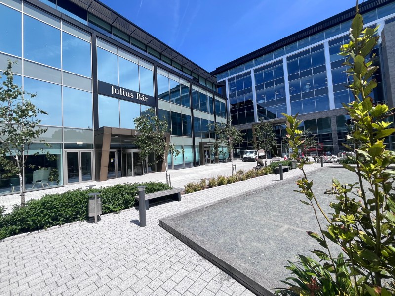 A modern office building with a sign saying Julius Baer. Trees and a square in front on a sunny day.