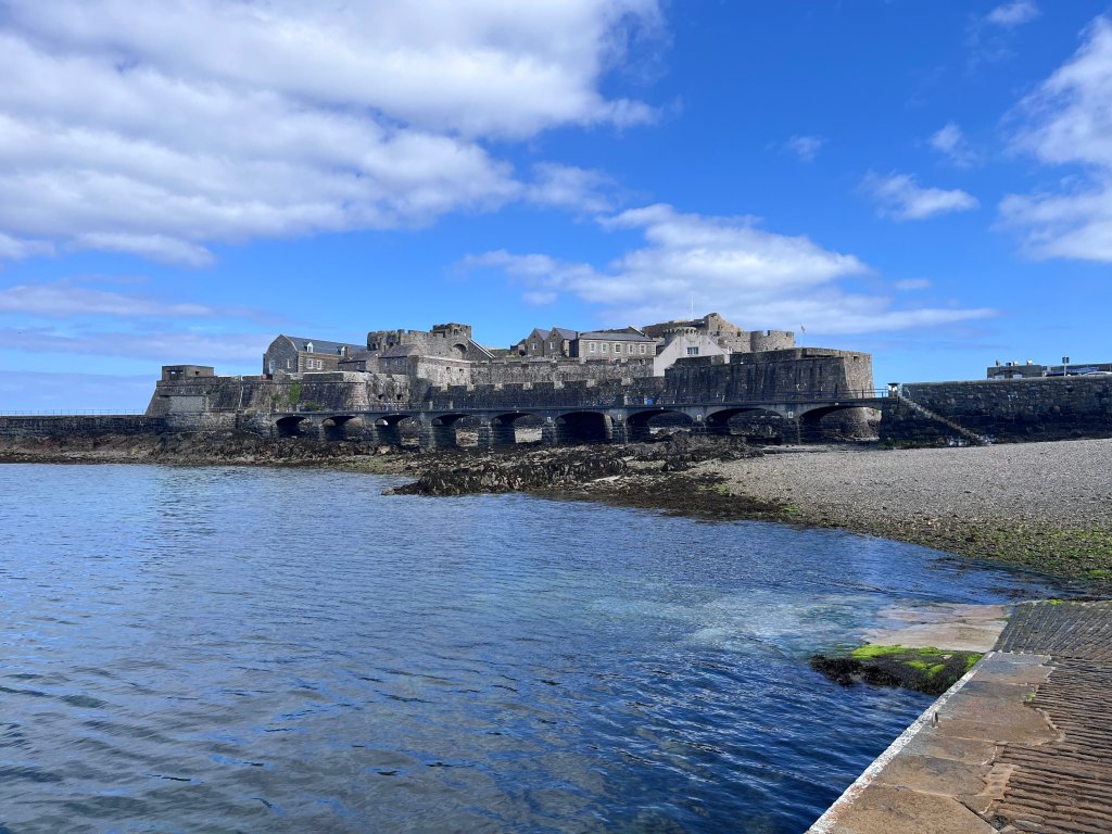 Wide view of Castle Cornet in Guernsey seen from the shoreline, with stone fortifications stretching along the water under a blue, partly cloudy sky.