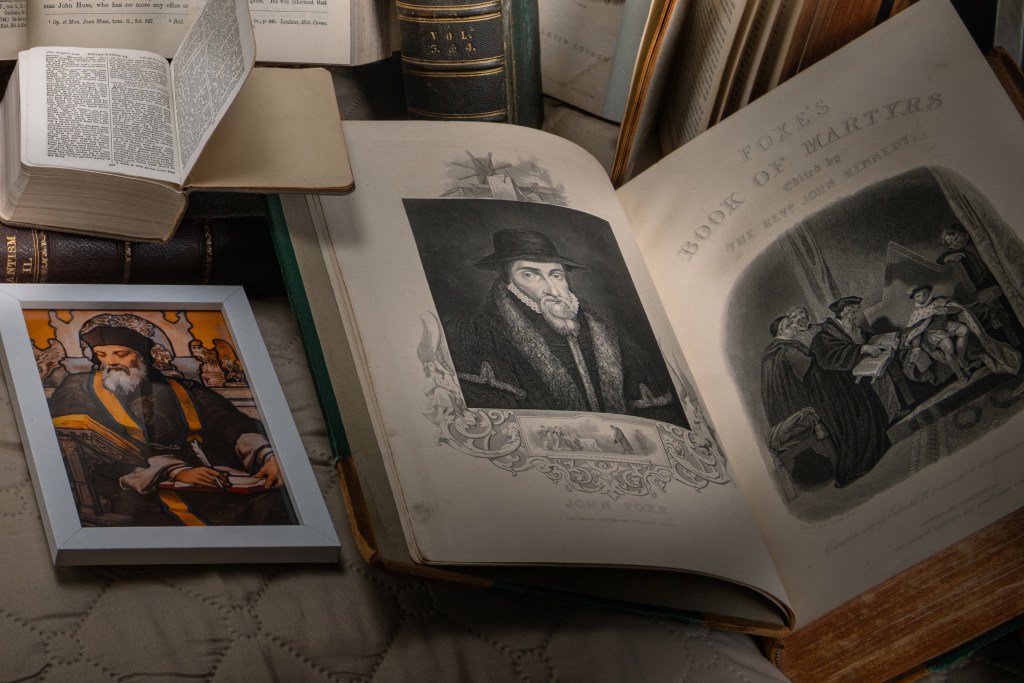 An open antique edition of Foxe’s Book of Martyrs lying on a fabric-covered surface, showing engraved portraits and a historical martyrdom scene, surrounded by other old books and a framed religious illustration.