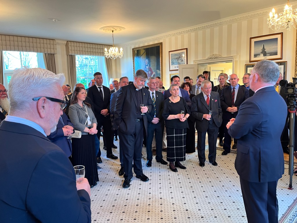 A group of people standing closely together in a formal reception room, listening to a man addressing them.