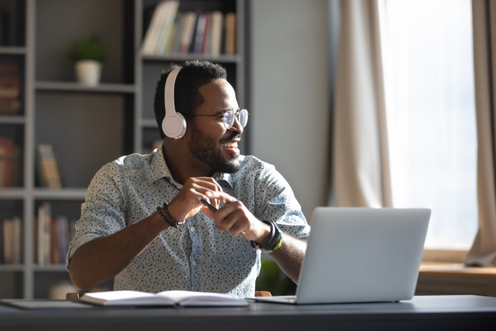 A man with glasses sat at a desk listening to something on headphones. He stares into the distance, with a laptop in front of him.
