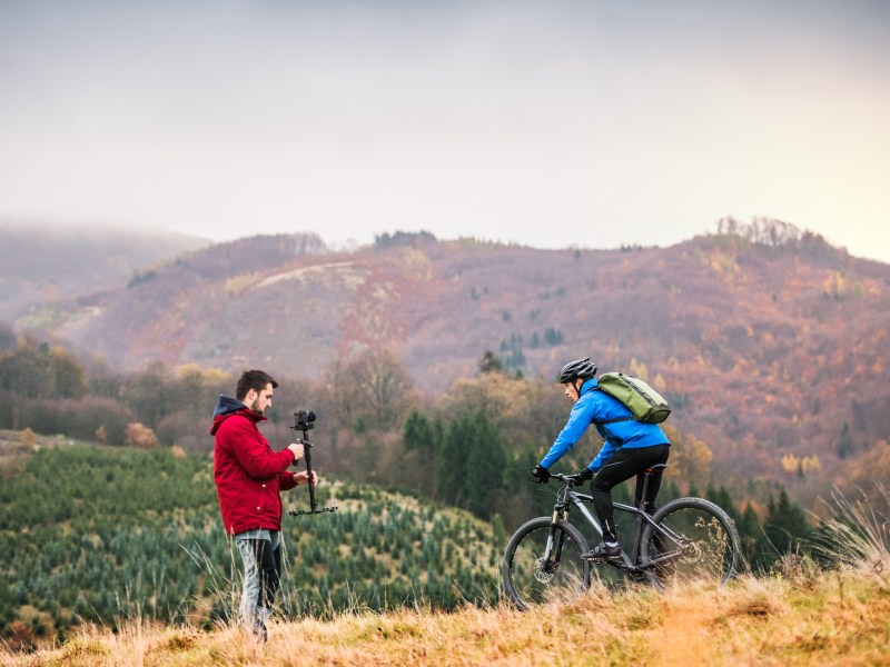 A filmmaker films a mountain biker in a field in a hilly area.