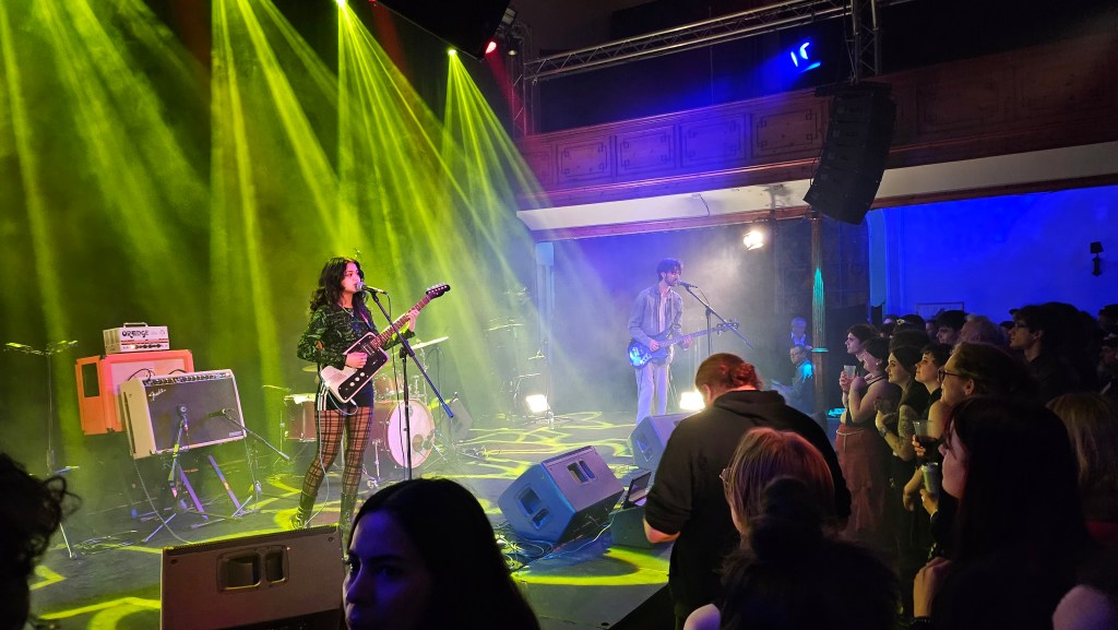 A female-fronted rock band, Hneyglaze, plays in front of an attentive crowd, with bright yellow and blue lights cutting through haze.