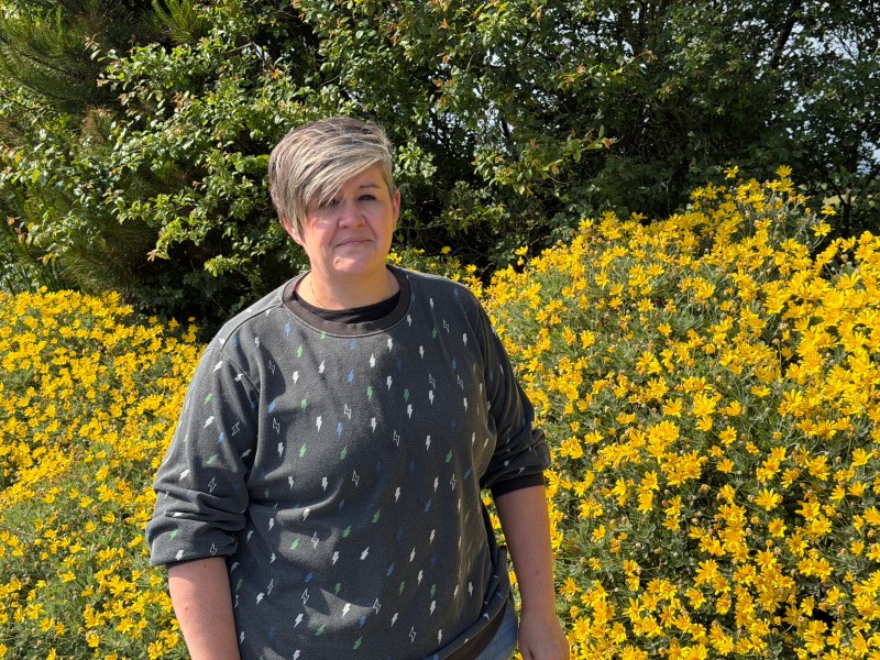 A woman with short grey hair stands in front of a bush full of yellow flowers on a sunny day.