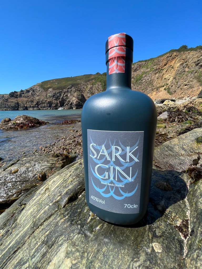 A bottle of Sark Gin on some granite rocks next to a pebble-strewn beach.