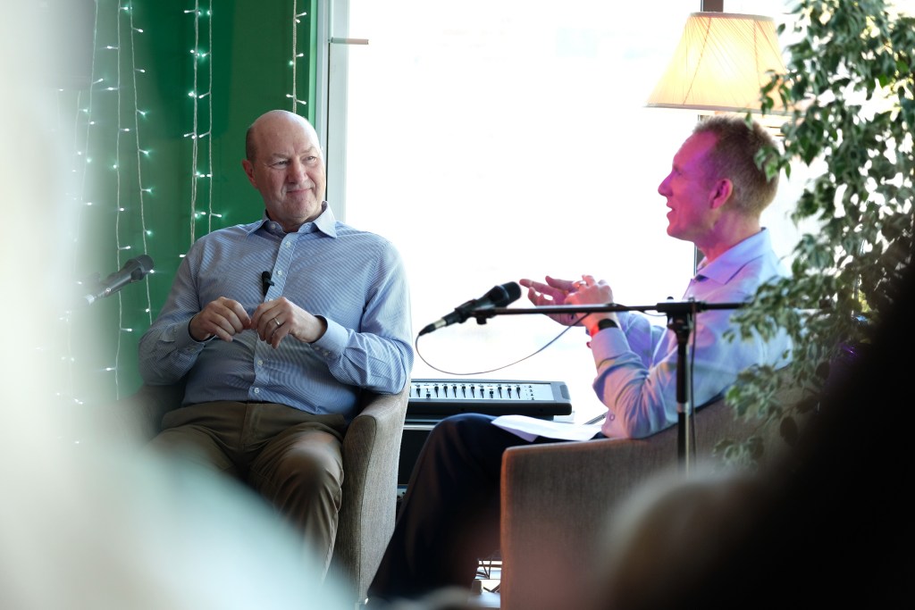 A bald man’s in a shirt sits and talks to another man, in comfy chairs with microphones in front of them.
