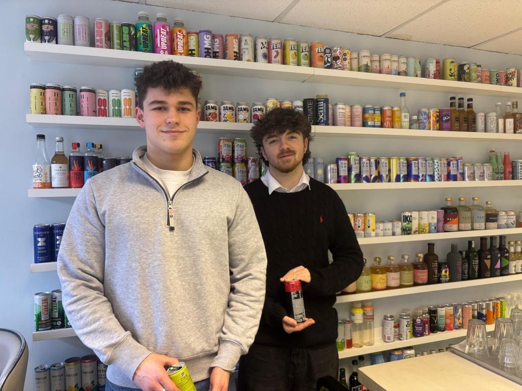 Two young med in a shop, with rows on drinks cans behind them.