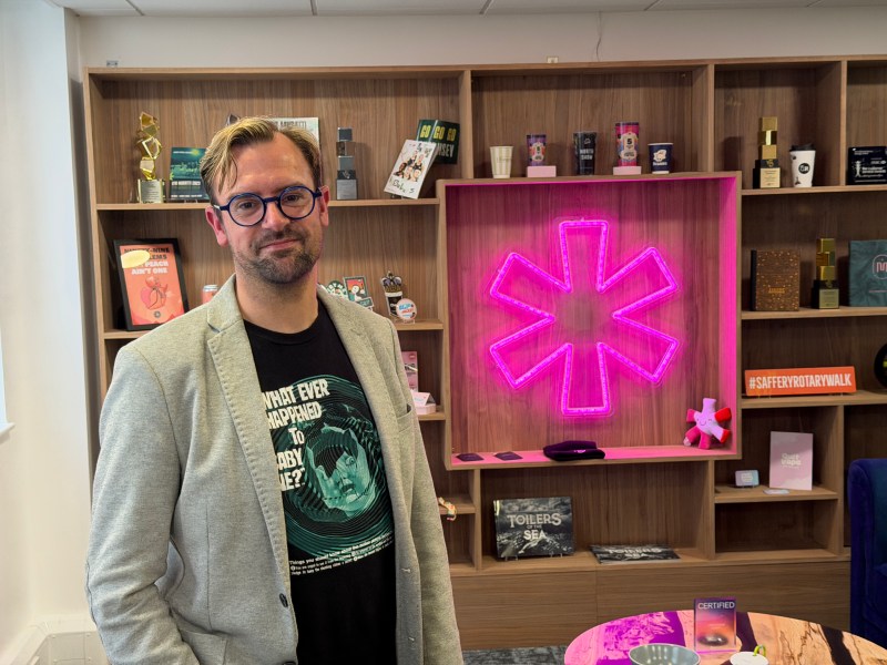 A man with glasses and stubble in a jacket and T-shirt stands smiling on a modern office with a neon logo.