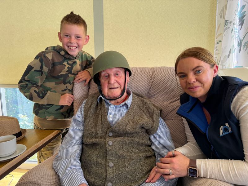 An old man in a chair, with a military-style helmet on. Next to him a smiling middle-aged woman and a small boy.