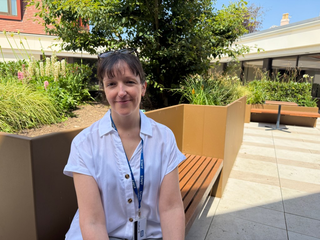 A smiling woman in a sunny school courtyard, with benches and trees.