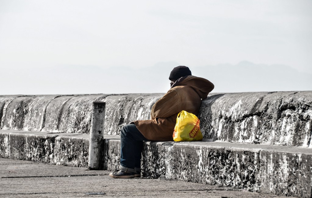 An ond man in a thick coat sits on a granite pier with a yellow carrier bag.