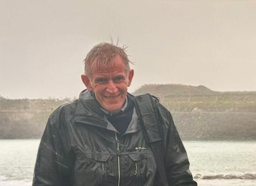 A man stands near the coast on a rainy day in a green coat. He's very wet.