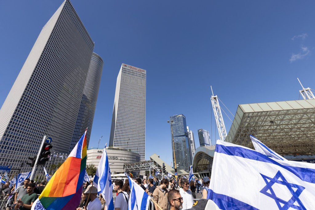 A demo in city surrounded by skyscrapers - featuring Israeli flags and a pride flag.