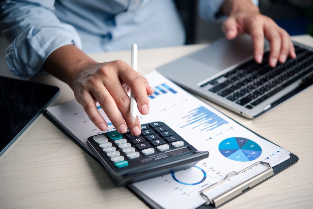 Calculator, finance Analyst presses calculator on budget report with charts laptop in office, reviewing data, accounting, numbers, analysis, planning, costs, and profit for decisions.
