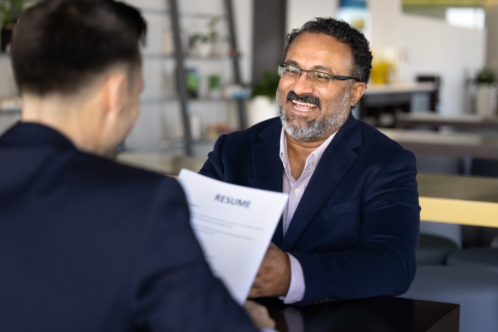 Asmiling middle-aged man in a suit hands his resume to an interviewer.