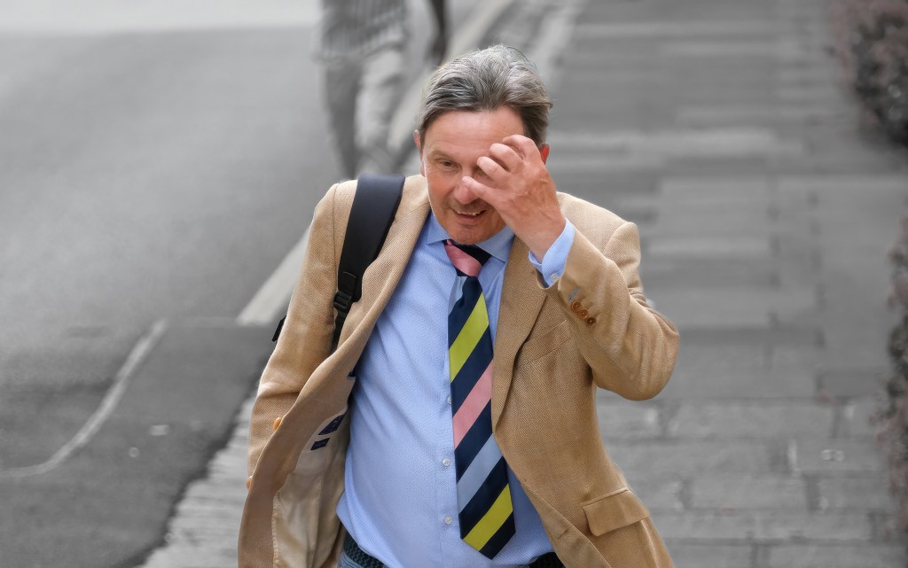 A man in a brown suit jacket puts his hand on his head while he walks up a street.
