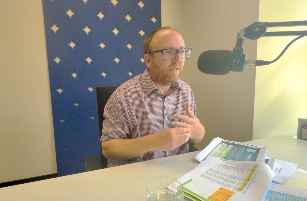 A balding man with a beard and glasses sits in front of a microphone.
